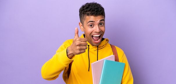 Jeune homme souriant en sweat jaune levant le pouce avec des cahiers, illustrant la confiance en soi et le syndrome du bon élève